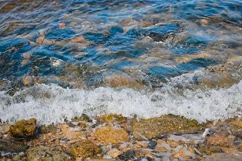 A small wave rolls on the seashore. Multi-colored stones underwater. Backgr.. Stock Photos