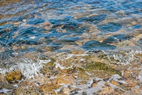 A small wave rolls on the seashore. Multi-colored stones underwater. Backgr.. Stock Photos