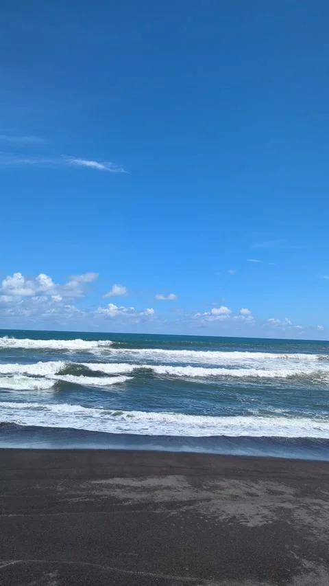 Small waves gently roll onto a dark sand beach. The deep blue sea meets a c.. Video stock 331179418