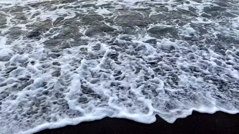 Small waves roll in one after another. Black volcanic sand beach. Video stock 141246931