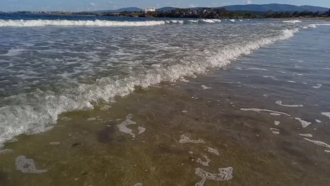 Small waves roll onto shore, mountains and resort town are visible on horizon. 스톡 동영상 266596455