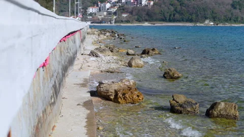 Small waves washing over rocks along a concrete seawall on a sunny coast 스톡 동영상 347181498