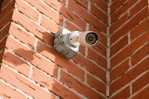Small white CCTV camera on the corner of the facade of brick building. Stock Photos