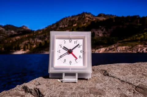 A small white clock with red hands sits on a rock by a body of water Foto stock