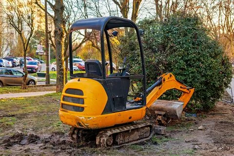 A small yellow excavator without a driver in front of a green bush. Stock Photos