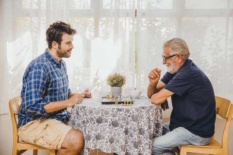 Smart elder playing chess board game at home care with younger man for stay b Stock Photos