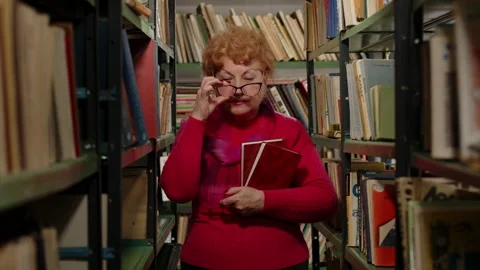Smart elderly teacher in glasses holding books in her hands in the library Stock Footage 170647893