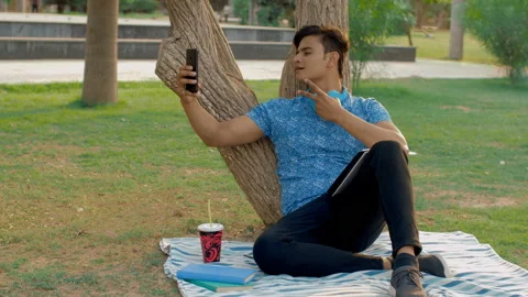 Smart Indian boy taking a selfie sitting in a beautiful park - modern technology Stock-Footage 154297180