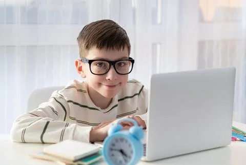 Smart nerd kid boy in glasses behind ear sitting at his desk at home using .. Stock Photos