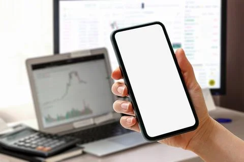 Smart phone with blank screen in hand in front of computers with financial ch Stock Photos