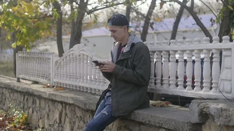 Smart young man sitting on a stone parapet and browsing the net in autumn 動画素材 119950394