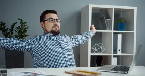 Smart young office worker in plaid shirt and glasses stretches while sitting at Stock Footage 147051536