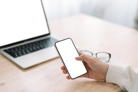 Smartphone with blank screen in hand in front of laptop with white screen on Stock Photos