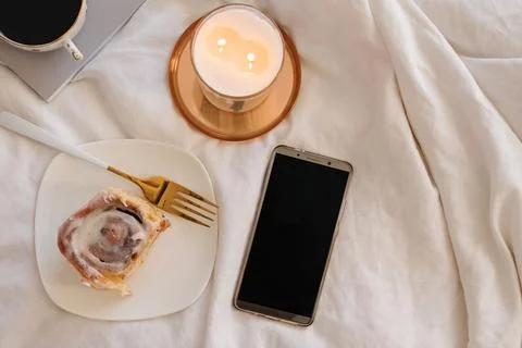 Smartphone with empty screen on the bed with cinnamon bun and burning candle Stock Photos