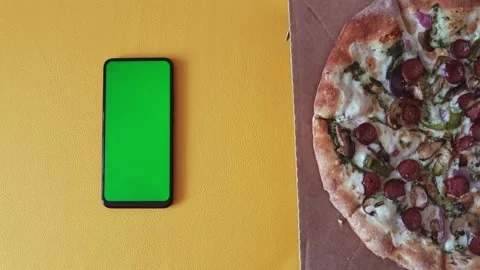 A smartphone with a green screen is lying on a yellow background a pizza appears Stock Footage 289231330
