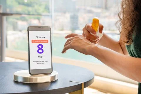 Smartphone with high UV alert in focus, while woman in background applies Stock Photos