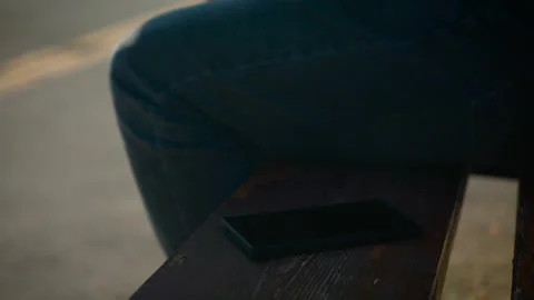 Smartphone left on wooden bench while person walking away in blurred background Stock Footage 333580353