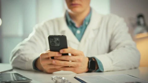 Smartphone in a physicians office glows as the young doctor sits typing a te Stock Footage 333831458