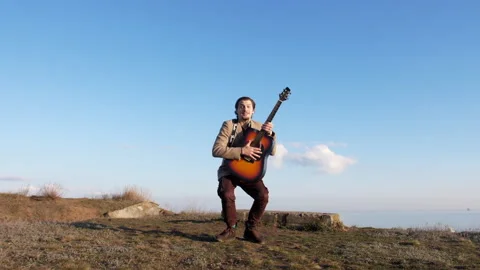 Smiley young guy jumping with guitar on the coast in sunny spring day Stock Footage 148941723