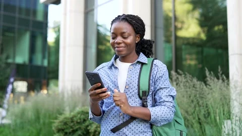 Smiling african american student with backpack checks her mobile phone while  Stock Footage 337056214