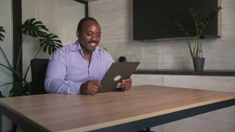 Smiling African man makes a video call using a tablet sitting at his desk at Stock Footage 201292459