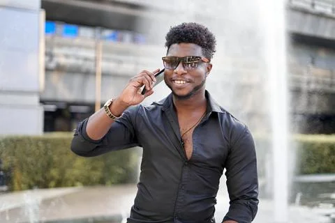 Smiling afro man talking to the mobile outdoors next to a fountain Stock Photos