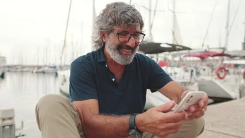 Smiling aged man sitting in port uses phone Stock Footage 253936637