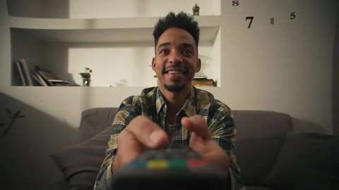 Smiling American man is sitting on couch with remote control and choosing movie. Video stock 302066796