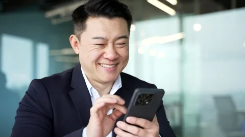 Smiling asian businessman is using mobile phone while sitting at a desk Stock Footage 321890188