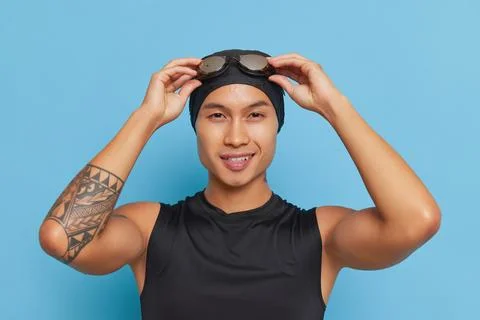 Smiling asian guy with diving goggles on his swimcap stands over blue background Stock Photos