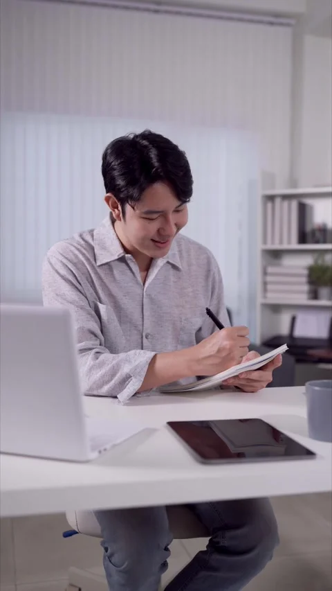 Smiling Asian man writing notes while video calling on laptop at home offic.. Stock Footage 345156603