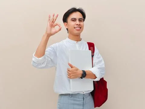 Smiling Asian student with backpack holding laptop and making ok sign. Stock Photos