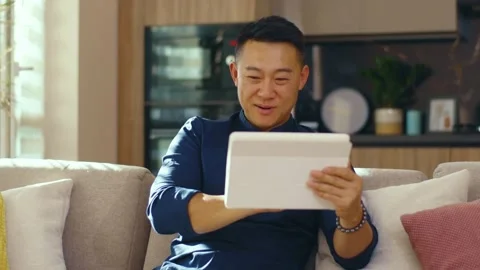 Smiling asian young man use tablet computer sitting on sofa at home. Feel happy Stock Footage 141006737