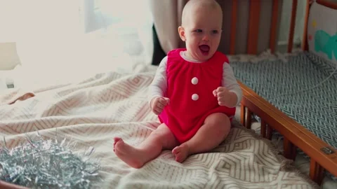 Smiling baby in a red romper waves and laughs while sitting on a bed in a bright Stock Footage 347686899