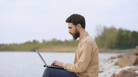 Smiling beard man working with laptop outdoors near the sea (or lake). Stock Footage 106834509