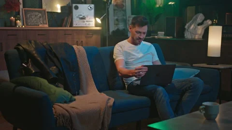 Smiling bearded good-looking man in white t-shirt sitting on sofa with laptop Stock Footage 232870255