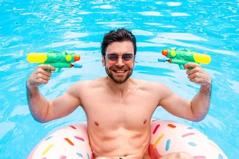 Smiling bearded guy posing with two toy water guns while using inflatable ring 스톡 사진
