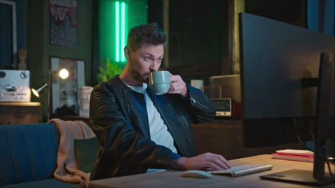 Smiling bearded man sitting in front of computer and drinking coffee in modern Stock Footage 244933755