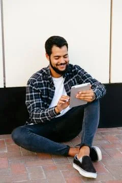Smiling bearded man using a computer tablet while sitting near the wall, outd Stock Photos