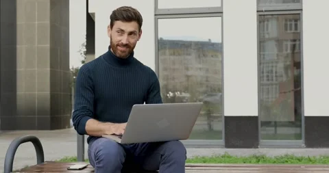 Smiling bearded man working on laptop while sitting on bench near modern Stock Footage 140680761