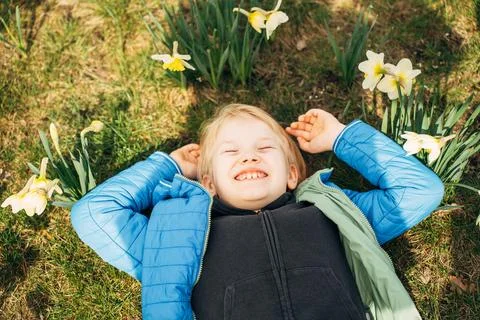 Smiling blond boy lying on green grass among blooming daffodils on a warm spring Stock Photos