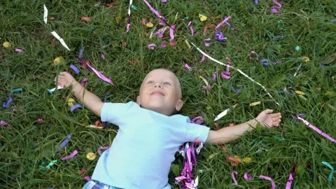 Smiling blond little boy lying on the green grass, looking up and waving at the 스톡 동영상 106679696