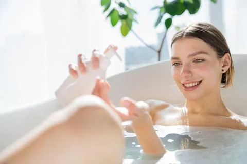 Smiling blondie using soap dispenser, while sitting in bath. Stock Photos