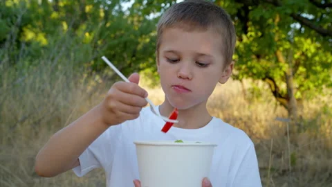 A smiling boy eats a vegetable salad on a paper plate outdoors. Stock Footage 311999013