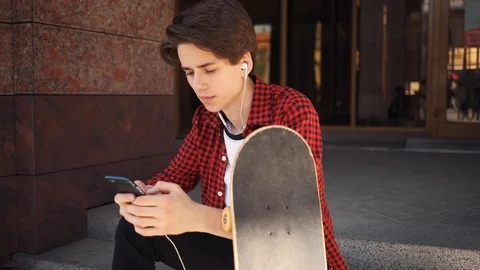 Smiling boy look at camera using mobile phone with headphones keeps skate guy Stock Footage 92051743