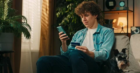 A smiling boy is sitting on the couch, he has made an online transfer for a new Stockbeeldmateriaal 218241743