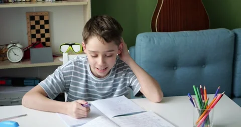 Smiling boy studying at home while reading textbook and writing notes in Stock Footage 345478047
