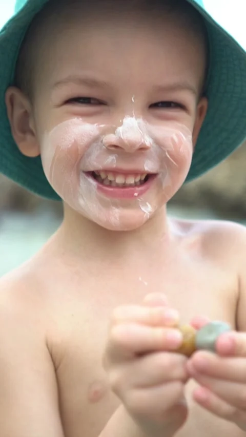 Smiling boy in a sun hat with sunscreen on his face looks at the camera and taps Stock Footage 347101808