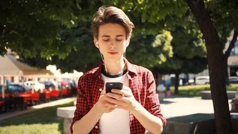 Smiling boy using mobile phone listens to music in headphones walk keeps skate Stock Footage 92051159