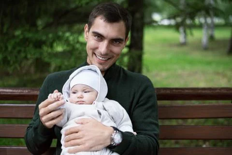 Smiling brunette dad in reen jacket sits on bench with baby in his arms in Pa Stock Photos